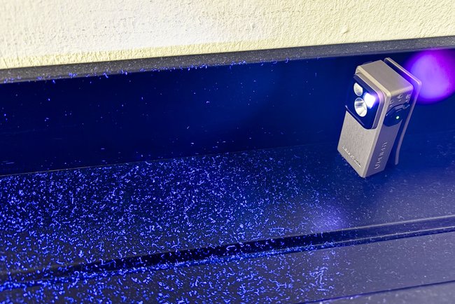 Die Mini-Taschenlampe Olight Oclip Ultra strahlt mit UV-Licht-Modus auf ein verstaubtes schwarzes Regal. Deutlich zu sehen sind Staubkörner, die jetzt blau aufleuchten und ansonsten kaum zu erkennen wären. Die Mini-Taschenlampe Olight Oclip Ultra strahlt mit UV-Licht-Modus auf ein verstaubtes schwarzes Regal. Deutlich zu sehen sind Staubkörner, die jetzt blau aufleuchten und ansonsten kaum zu erkennen wären.