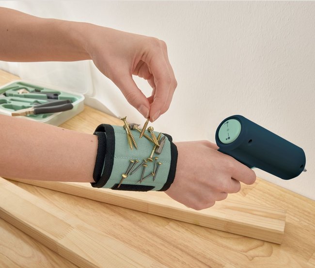 Magnetarmband am Handgelenk einer Person, an dem mehrere Nägel und Schrauben hängen. Die Person greift mit der anderen Hand nach einer Schraube. Magnetarmband am Handgelenk einer Person, an dem mehrere Nägel und Schrauben hängen. Die Person greift mit der anderen Hand nach einer Schraube.