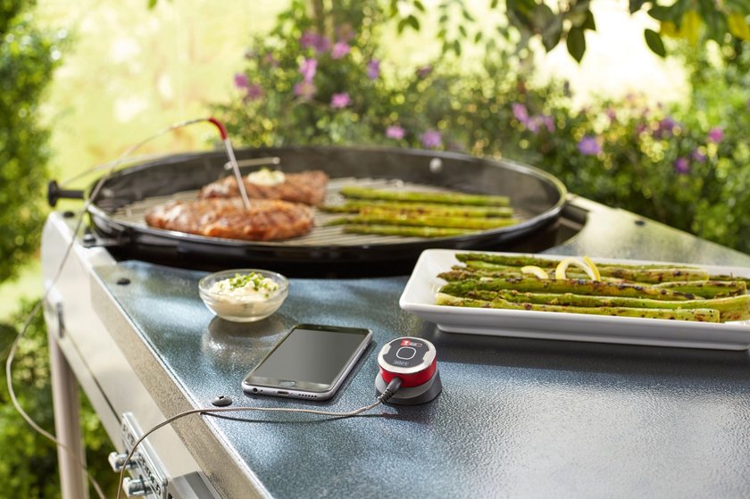 Ein Grill mit zwei Steaks und Spargel darauf. Ein Fleischthermometer ist in eines der Steaks gesteckt und mit einem Kabel mit einem Grillthermometer verbunden, das neben dem Smartphone liegt. Ein Grill mit zwei Steaks und Spargel darauf. Ein Fleischthermometer ist in eines der Steaks gesteckt und mit einem Kabel mit einem Grillthermometer verbunden, das neben dem Smartphone liegt.