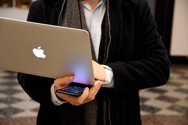 Eine Person in schwarzem Anzug trägt ein geöffnetes Macbook und hält ein iPhone in der Hand.