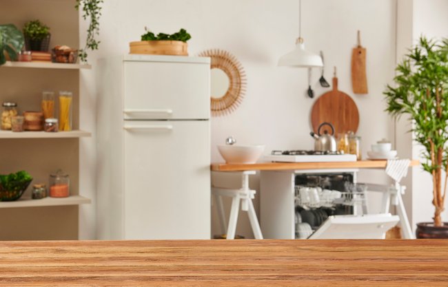 Eine Küche im Überblick: Im Vordergrund ist ein Esstisch aus Holztisch und im Hintergrund nebeneinander ein Küchenregal, ein Kühlschrank und eine Spülmaschine.