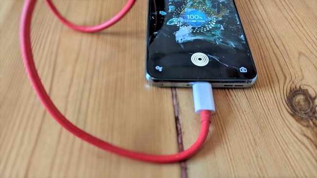 Auf einem Dielenboden wird das Smartphone OnePlus 10T mit einem roten Ladekabel aufgeladen.