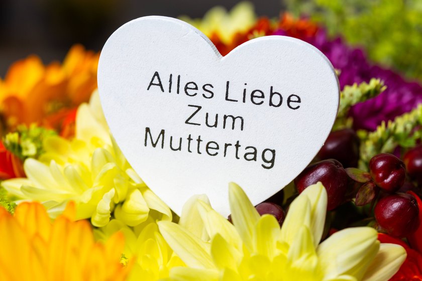 Herzförmiges Schild inmitten von Blumensträußen mit der Aufschrift "Alles Liebe zum Muttertag". 