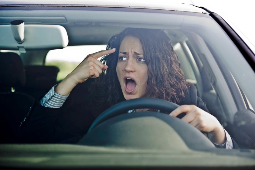 Frau schaut wütend aus einem Auto und tippt sich mit dem Finger an die Stirn. 