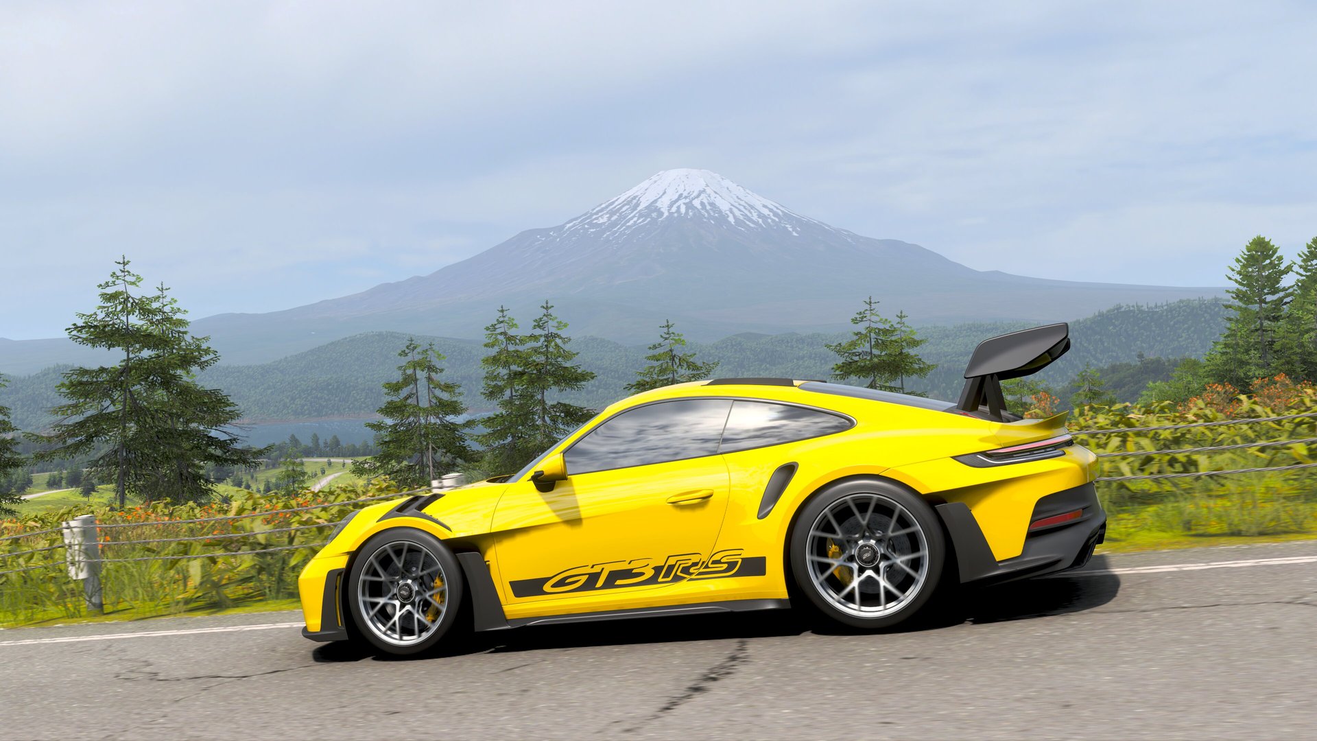 A yellow Porsche GT3 RS drives on an asphalt road with a snow-capped mountain in the background. The mountain is surrounded by green hills and trees, and some coniferous trees can be seen in the foreground.