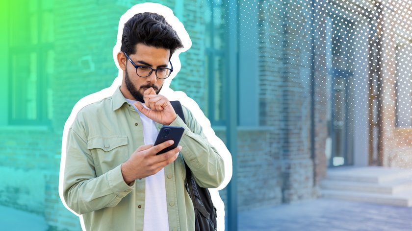 Junger Mann mit Brille schaut nachdenklich auf sein Smartphone. Im Hintergrund ist eine Häuserecke zu sehen.