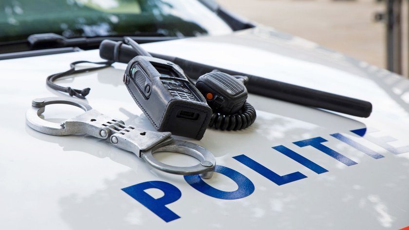 Nahaufnahme von Handschellen, einem Funkgerät und einem Schlagstock, die auf der Motorhaube eines Polizeifahrzeugs liegen. Auf der Motorhaube ist der Schriftzug "Politie" teilweise sichtbar.  