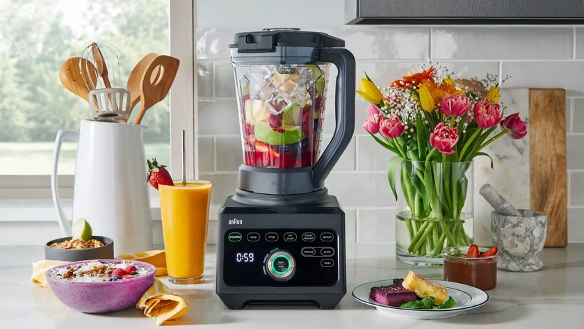 Ein Braun PowerBlend 9 Mixer steht auf einer Küchenarbeitsfläche. Der Behälter ist mit frischem Obst, darunter Beeren, Äpfel und Kiwi, sowie Eiswürfeln gefüllt. Auf der Arbeitsfläche befinden sich ein Glas mit gelbem Smoothie, eine violette Smoothie-Bowl, ein Teller mit Dessert, ein Glas mit Schokoladencreme und eine Vase mit bunten Tulpen. Im Hintergrund stehen Küchenutensilien in einem weißen Behälter und ein Mörser aus Stein.  