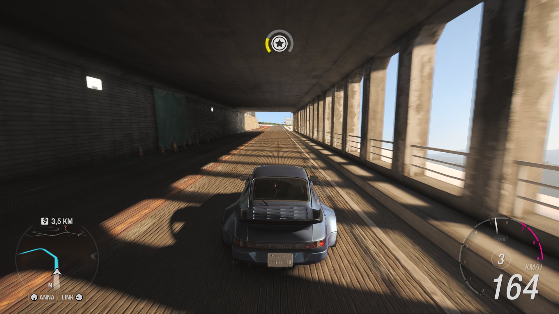 Rear view of a blue Porsche sports car driving on a wooden surface in a tunnel. Sunlight shines through the open sides of the tunnel, casting long shadows. A speedometer showing 100 mph (164 km/h) can be seen on the dashboard.
