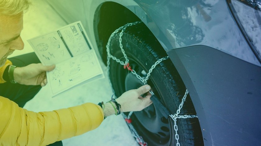 Ein Mann prüft die Schneeketten an seinem Auto mit Bedienungsanleitung in der Hand.