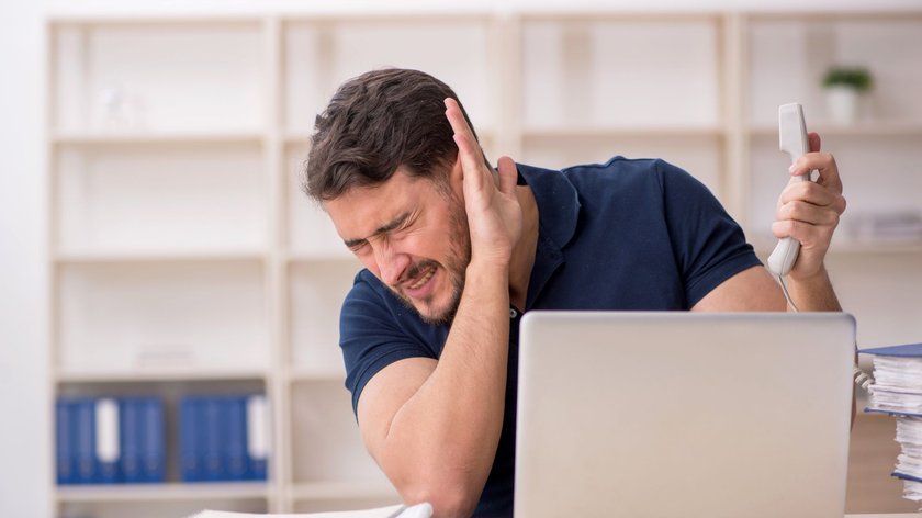 Mann am Schreibtisch hält sich vor Schmerz das Ohr zu, während er einen Telefonhörer in der anderen Hand hält. Vor ihm steht ein Laptop, daneben liegen Dokumente und ein Stapel Papier.