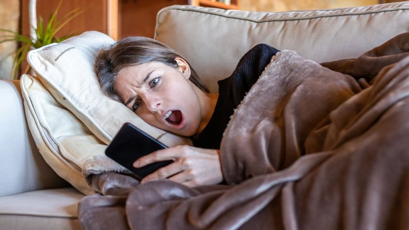 Junge Frau liegt auf Sofa, guckt aufs Handy und macht ein überraschtes Gesicht.