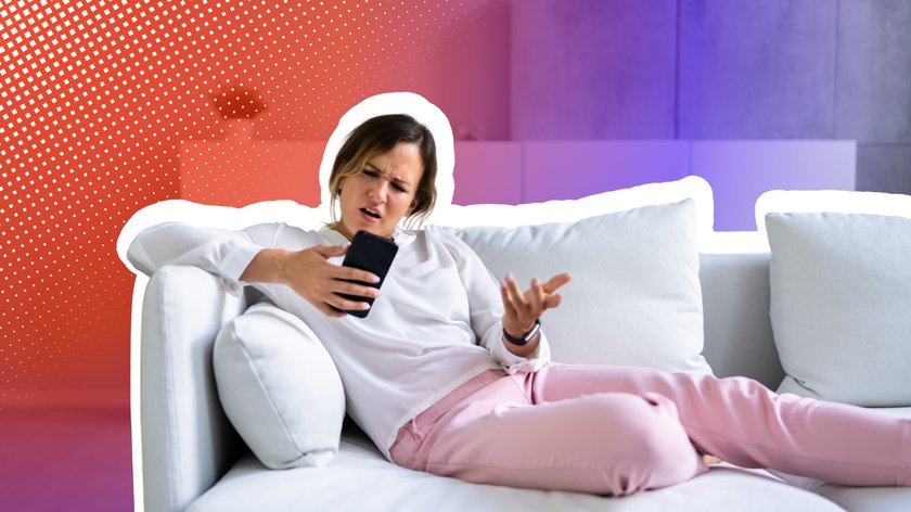 Frau in weißem Oberteil und rosa Hose sitzt auf einem weißen Sofa und blickt verärgert auf ihr Smartphone. Sie hält das Telefon in einer Hand und gestikuliert frustriert mit der anderen. Der Hintergrund ist mit einem bunten Farbverlauf in Rot und Violett sowie einem grafischen Punktmuster gestaltet.