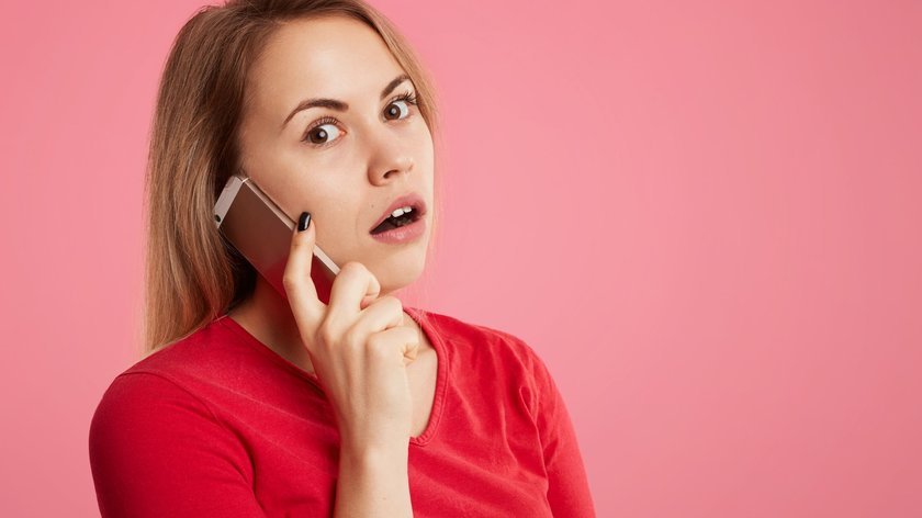 Schockierte Frau erhält Telefonanruf, spricht mit Fremden, hält Smartphone in der Hand und blickt mit überraschtem, verängstigtem Gesichtsausdruck in die Kamera.