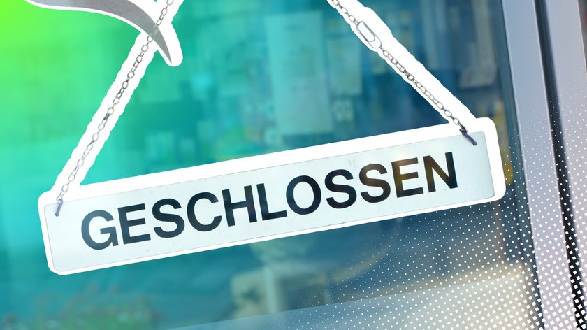 Schild mit der Aufschrift "GESCHLOSSEN" hängt an einer Kette vor einem Schaufenster. Der Hintergrund ist unscharf, mit blauen und grünen Farbtönen.