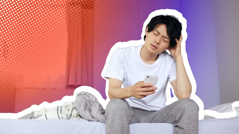 Junger asiatischer Mann sitzt auf dem Bett, schaut fragend auf sein Handy und kratzt sich dabei am Kopf.