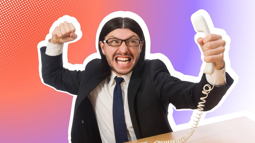 Ein Mann mit langen, dunklen Haaren, Brille und Bart trägt einen schwarzen Anzug mit weißem Hemd und dunkelblauer Krawatte. Er hält wütend einen Telefonhörer in der Hand, während er seine Faust ballt und schreit.
