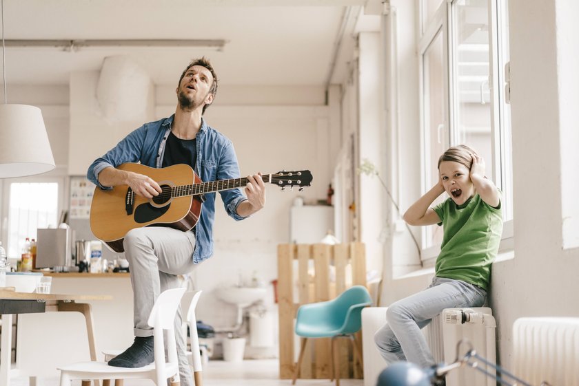 Mann spielt Gitarre, Kind hält sich genervt die Ohren zu. 