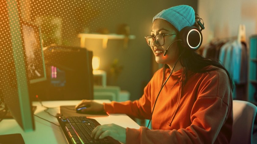 Frau mit Brille und Kopfhörer mit Mikrofon sitzt vor einem Computerbildschirm und tippt auf einer beleuchteten Tastatur. Sie trägt einen orangefarbenen Hoodie und eine Mütze. Der Raum ist dezent beleuchtet, im Hintergrund sind Regale und Kleider zu sehen.