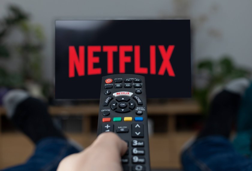 Hand hält Fernbedienung, in der Ferne ist Fernseher mit Netflix-Logo