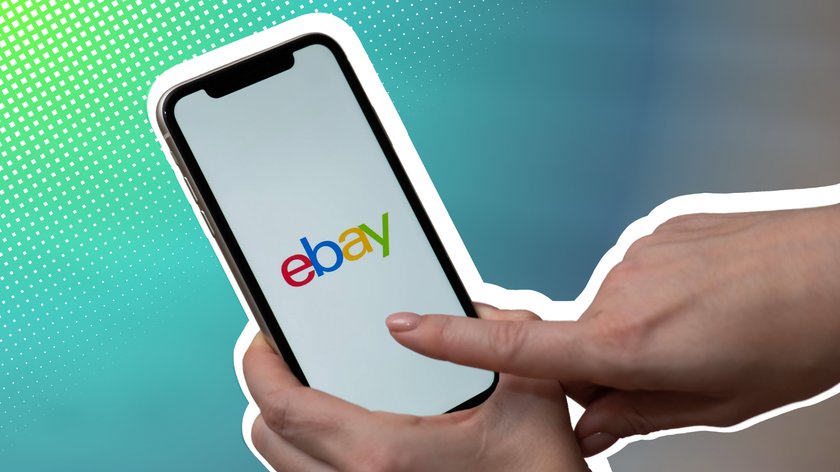 Nahaufnahme einer Hand, die ein Smartphone hält, auf dessen Bildschirm das bunte eBay-Logo zu sehen ist. Eine zweite Hand zeigt mit dem Finger auf den Bildschirm.