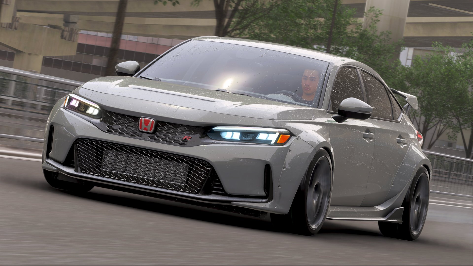 A gray Honda Civic Type R drives on a wet road. The car has an aggressive front grille with a red Honda logo and sharp headlights. Buildings and trees can be seen in the background.
