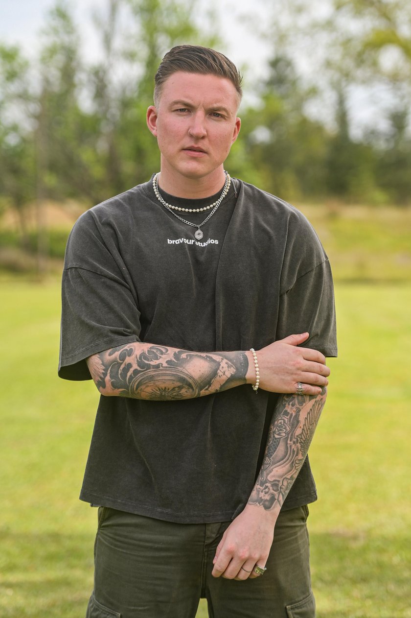 Ein junger Mann mit tätowierten Armen, der ein schwarzes T-Shirt trägt, steht vor einer grünen Wiese mit Bäumen im Hintergund.