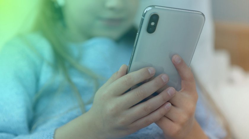 Ein Kind mit hellblauem Pullover hält ein älteres Modell eines iPhones vor sich in der Hand.