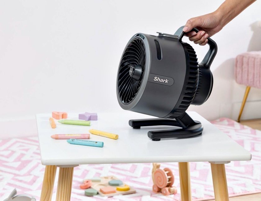 Ein schwarzer Shark FlexBreeze HydroGo Ventilator mit Sprühnebel steht auf einem weissen Tisch mit Holzbeinen. Eine Hand greift den Tragegriff des Ventilators. Auf dem Tisch liegen bunte Kreiden und Spielzeug. Im Hintergrund sind ein rosa gemustertes Teppichmuster, ein Kinderstuhl und weitere Spielzeuge zu sehen.