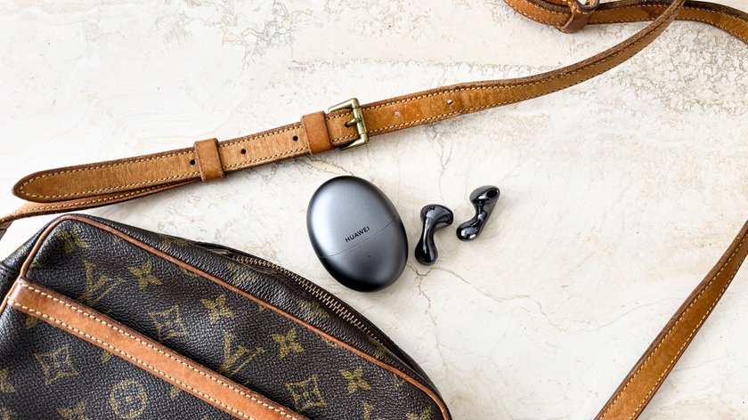 Die Huawei FreeBuds 6 in Schwarz, bestehend aus einem ovalen Ladecase mit Huawei-Logo und zwei kabellosen Ohrhörern, liegen auf einer hellen, marmorierten Oberfläche. Im Hintergrund sind ein brauner Lederriemen und eine Louis Vuitton-Tasche mit braunen Akzenten zu sehen.  