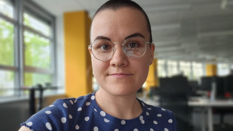 Eine Frau mit kurzen dunklen Haaren, Brille und blauem Shirt mit weißen Punkten schaut in die Kamera.