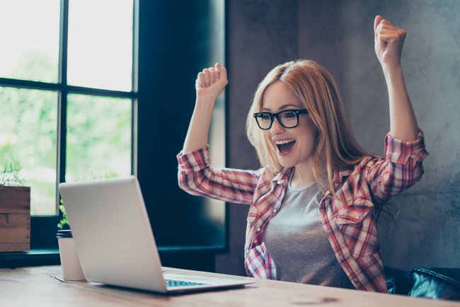 Work complete. Young, blonde, specialist rejoicing her finished work with raised fists sitting at her desk