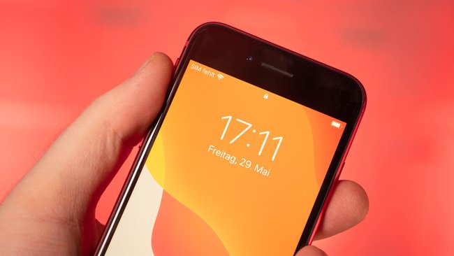 Vor einem roten Hintergrund hält eine Hand ein Smartphone. Das Modell ist ein iPhone SE 2022.