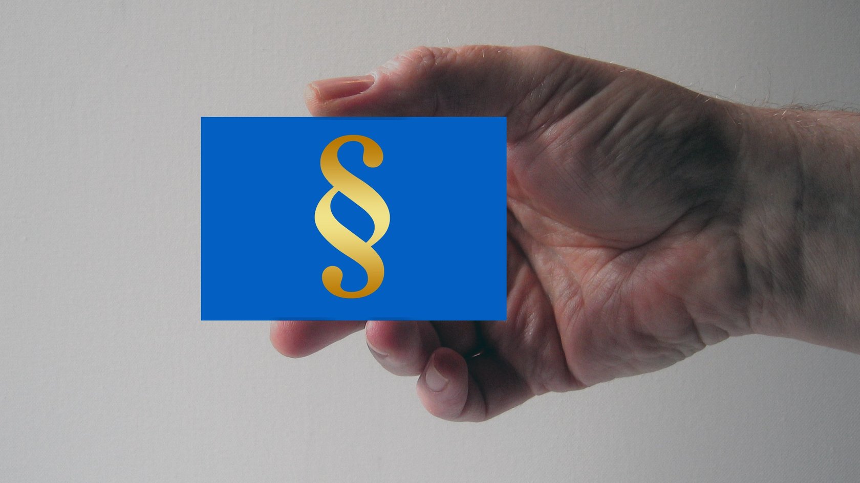Eine Hand hält eine blaue Karte, die ein goldenes Paragraphenzeichen ( § ) zeigt. Der Hintergrund ist einfarbig hell.  