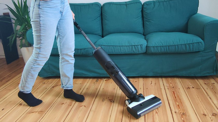 Im Vordergrund sieht man eine Person mit hellblauen Jeans, die einen schwarz-weißen Akku-Staubsauger in der Hand hält. Im Hintergrund steht ein grünes Sofa.