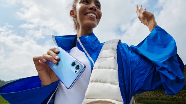 A woman wearing a blue and white jacket is holding the light blue Oppo Find X5 Lite smartphone.