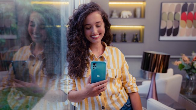 A woman with long brown hair in a living room looks at the blue Moto G22 smartphone in her hand.