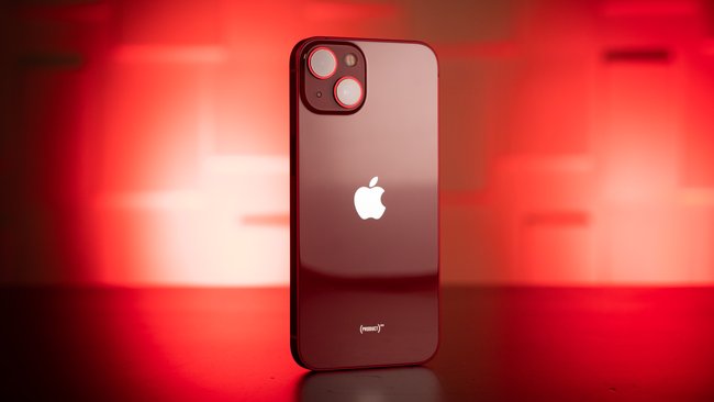 Im Vordergrund sieht man die Rückseite eines schwarzen iPhone 13, das senkrecht auf einer Tischplatte steht. Das Smartphone wird rot angestrahlt.