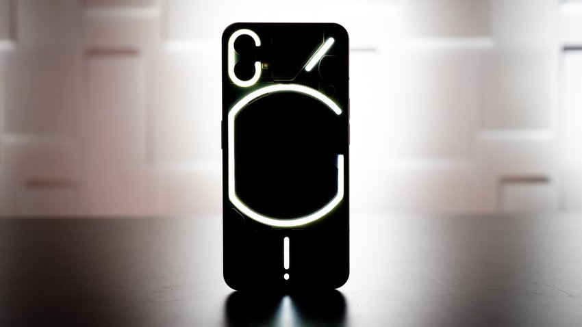 Im Vordergrund steht ein schwarzes Smartphone mit weißen Lichtstreifen auf der Rückseite. Die Unterlage ist schwarz und das Handy wird mit einem weißen Licht angestrahlt.