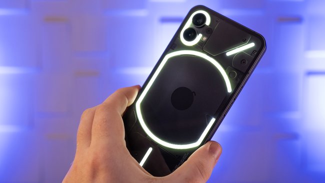 Eine Hand hält das Smartphone Nothing Phone (1). Die Rückseite des Handys hat ein leuchtendes Muster. Im Hintergrund ist violettes Licht zu sehen.