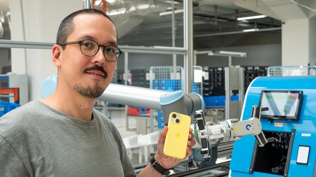 Stefan steht mit einem iPhone in der Hand vor einem Prüf-Roboter von Rebuy