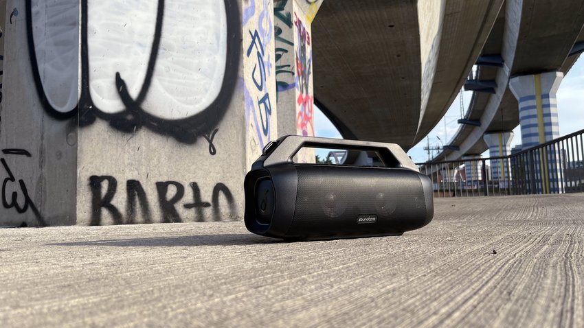Ein tragbarer Bluetooth-Lautsprecher mit dem Modellnamen Anker Soundcore Motion Boom Plus steht im Freien auf dem Boden. Die Sonne scheint, im Hintergrund sind Graffiti an einer Wand zu sehen.