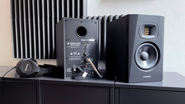 Rückseite und Vorderseite des Studiomonitors Adam Audio T5V. Links daneben zum Größenvergleich ein Over-Ear-Kopfhörer von Shure.