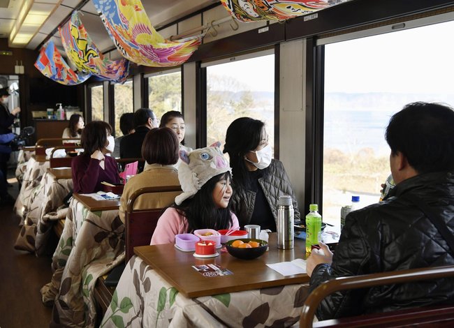 In einem Zug sitzen Menschen an kleinen Heiztischen namens Kotatsu. Die Decken sind mit Pflanzen verziert.