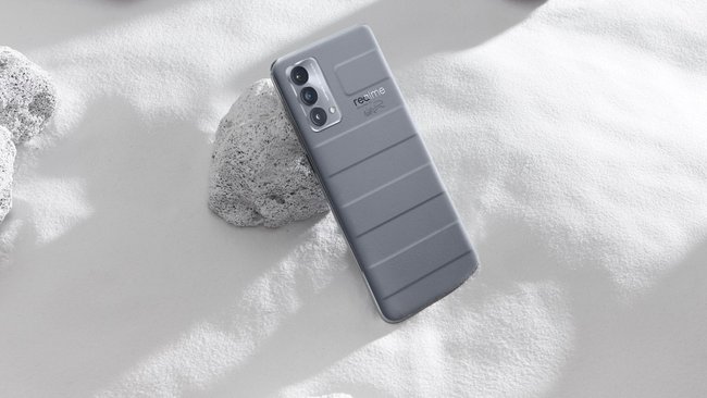 Das graue Smartphone Realme GT Master Edition wird im Sand von einem Stein gestützt.