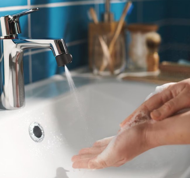 In einem blauen Badezimmer ist im Vordergrund ein Waschbecken mit aufgedrehtem Wasserhahn. Zwei Hände werden mit Seife gewaschen.