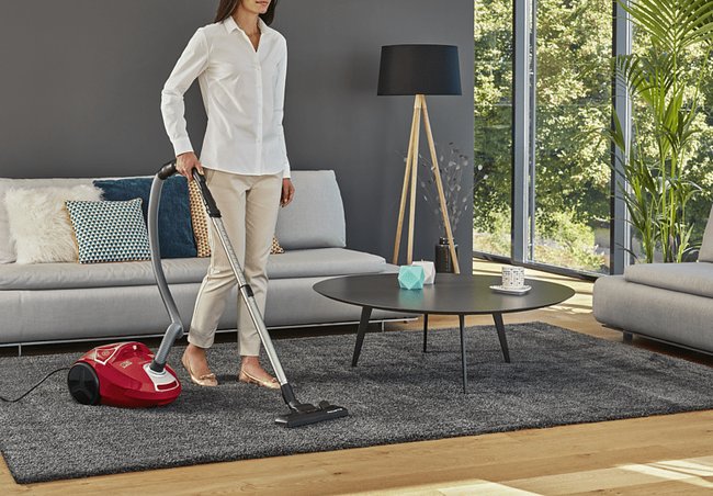 Eine weiblich gelesene Person saugt im Wohnzimmer mit einem roten Staubsauger den Boden.