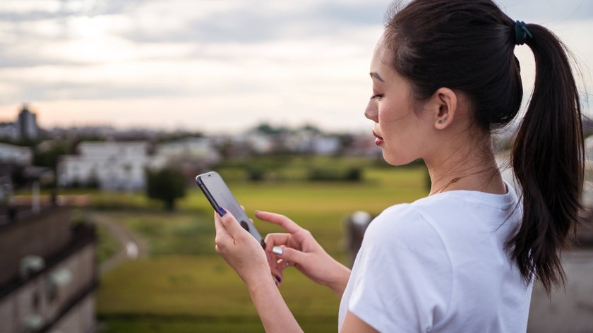 Eine weiblich gelesene Person mit weißem T-Shirt steht an einer Wiese und schaut auf ihr Handy.