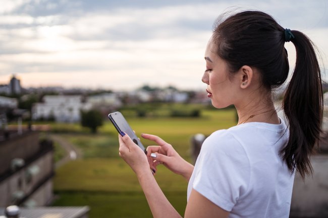 Eine weiblich gelesene Person mit weißem T-Shirt steht an einer Wiese und schaut auf ihr Handy.