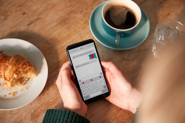 Eine Person sitzt an einem Tisch, vor ihr Kaffee und Croissant. Sie hält das Handy in der Hand mit einem romantischen Chat.